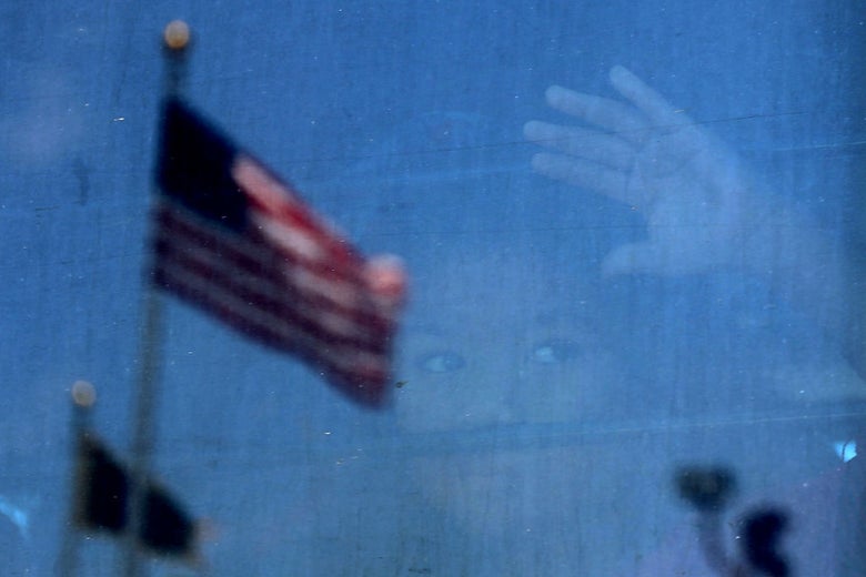 A young child's face can be seen through the window of a bus against which a United States flag is reflected.